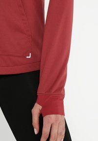 Giacca sportiva rossa con maniche lunghe, caratterizzata da una texture liscia, aperture per il pollice e una tasca discreta sul lato, abbinata a leggings neri.