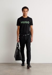 Fekete póló zöld "HUGO" felirattal, fekete nadrág és fekete cipő. A modell egy fekete vászontáskát tart, minimalista háttér előtt áll.