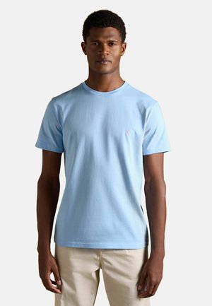 Man wearing light blue short-sleeve crew neck t-shirt and beige pants, standing against a plain white background.