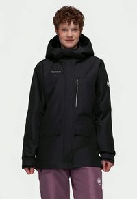 Junge Person mit kurzen lockigen Haaren, die eine schwarze Mammut-Jacke mit Kapuze und mauvefarbene Hosen trägt, steht vor einem schlichten Hintergrund.