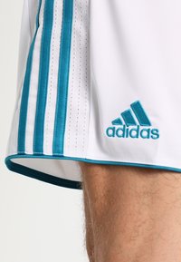 Adidaksen urheilushortsit, valkoiset tehosteilla teal-värillä. Sivuille on ommeltu kolme teal-väristä raitaa ja teekaudalla brodeerattu logo. Kevyt kangas.