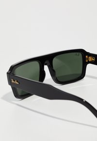 Ochelari de soare rectanguli negri cu lentile verzi. Caracteristici accente aurii pe cadru și logo-ul Ray-Ban pe ramă. Textură mată pe întreaga suprafață.