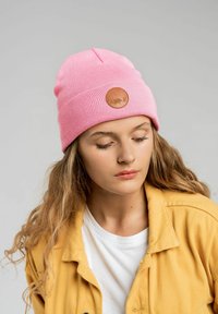 Różowa, prążkowana czapka z klapką ze skórzanym naszywkiem z przedstawieniem zwierzęcia, noszona z żółtą kurtką na białym t-shircie.