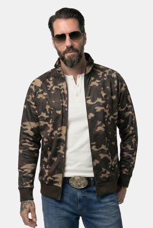 Hombre con chaqueta de camuflaje, camiseta henley blanca, vaqueros azules, gafas de sol y una hebilla de cinturón grande, de pie con las manos a los lados.