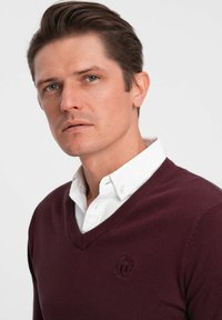 Marin v-nacksweater i mjukstickat material, med ett broderat logotyp på bröstet, bärs över en vit skjorta med knappar.