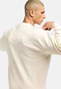 Beige pulóver bordás mandzsettákkal, dombornyomott szöveggel és grafikai tervezéssel a hátsó oldalon. Puha, textúrált anyag; laza szabású.