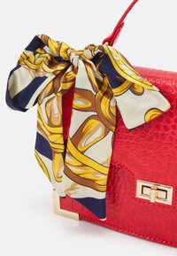 Sac à main en cuir rouge avec une finition texturée, orné d'un foulard en soie décoratif aux motifs dorés et bleu marine noué à la poignée. Détails en métal doré.