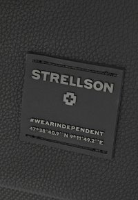 Materiale nero testurizzato con un'etichetta simile alla gomma che mostra "STRELLSON", un simbolo di croce e l'hashtag #WEARINDEPENDENT, con le coordinate sottostanti.