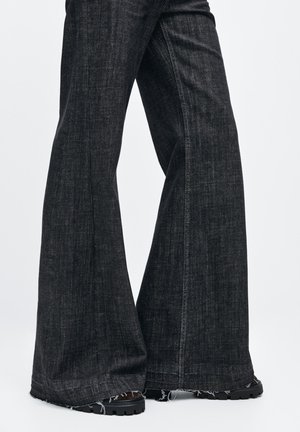 Zwarte flare jeans met een gestructureerde afwerking, voorzien van rafelige zoom en contrasterende stiksels. Het materiaal is denim met een lichte glans.