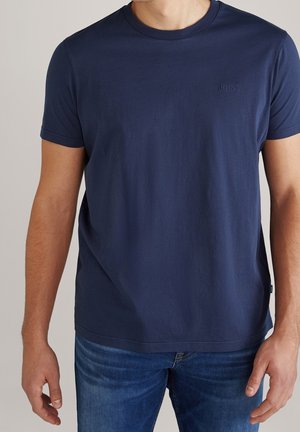 Mężczyzna ubrany w prosty granatowy krótki rękaw i niebieskie dżinsy, stojący na neutralnym tle.