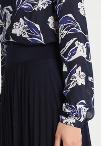 Blusa floreale con design blu navy e bianco, maniche lunghe e vita elastica; abbinata a una gonna plissettata di colore blu navy.