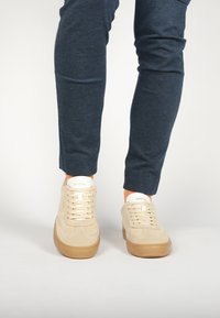 Beige mokasiniai sportbačiai su suapvalinta forma, rusvos gumos padas ir baltos juostos. Kulšnyje matosi jogger stiliaus tamsiai mėlynos kelnės su tekstūra.