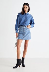 Denim shirt with studded accents, long sleeves, and button closure, paired with a denim skirt featuring front buttons and a fitted silhouette. Black ankle boots complete the look.