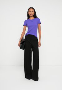 Donna che indossa una maglietta aderente viola, pantaloni neri a gamba larga, tiene una grande pochette nera, in piedi contro uno sfondo bianco.