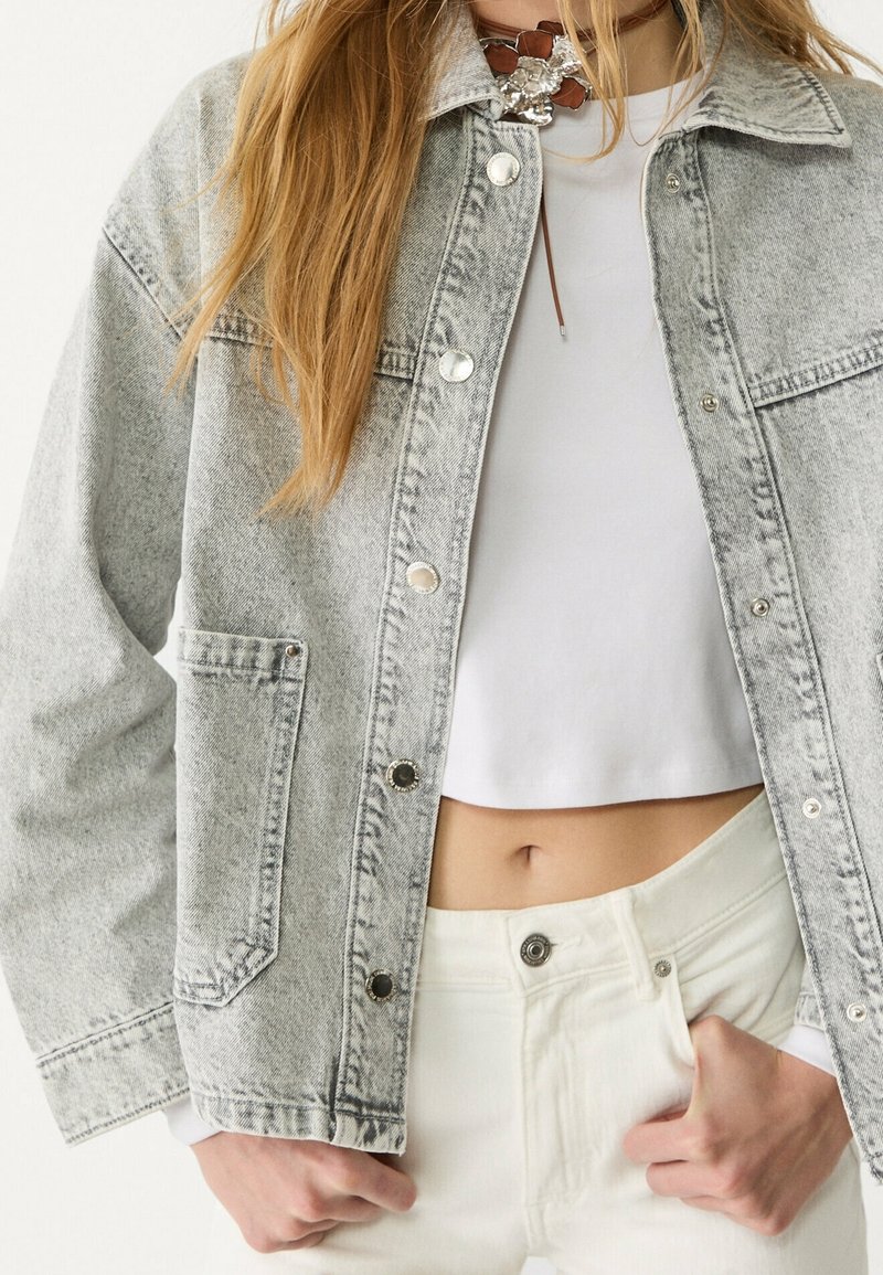 Žena v světle šedé džínsové bund�ě přes bílé crop top a bílé kalhoty s vysokým pasem, s koženým náhrdelníkem kolem krku.