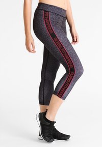 Svarta capri leggings med ett geometriskt mönster, som har en röd grafisk textdesign längs de yttre sömmarna. Tillverkade av ett elastiskt, lätt material.