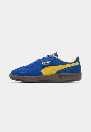 Sneakers in suede blu con accenti gialli e suola in gomma marrone scuro. Presentano un design con lacci e un logo del marchio in rilievo sul lato.