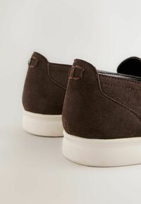Chaussures à enfiler en daim marron avec semelles en caoutchouc blanches, couture arrière visible, vues de dos sur un fond clair.