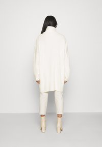 Överdimensionerad vit stickad tröja med hög krage och ribbat mönster, kombinerad med krämfärgade byxor och beige klackade ankelstövlar.