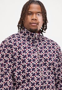 Veste zippée avec un motif géométrique en bordeaux, blanc et bleu marine. Col montant et texture de tissu lisse. Détail de fermeture éclair en argent.