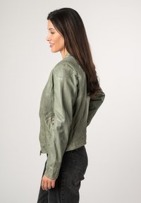 Giacca verde simile alla pelle con un design aderente, chiusura con zip e maniche lunghe. Abbinata a jeans grigi, che mostrano una texture liscia e un taglio moderno.