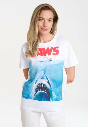 White cotton t-shirt featuring a graphic of a shark and swimmer with "JAWS" text in bold red. Short sleeves and relaxed fit.