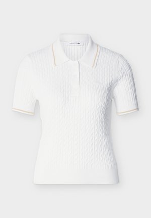 Polo blanc à manches courtes en maille torsadée, avec une bordure beige sur le col et les manches, une patte de boutonnage, et le logo Lacoste sur la poitrine.