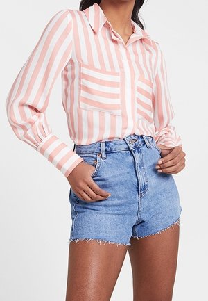 Kobieta ubrana w różowo-białą koszulę z pionowymi paskami na guziki, z dwoma przednimi kieszeniami, oraz wysokim stanem niebieskie szorty jeansowe z postrzępionymi brzegami.