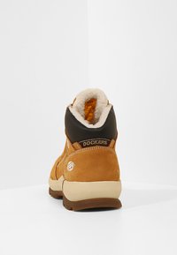 Botte chelsea en daim marron avec doublure en fausse fourrure blanche, accent noir à l'arrière et marquage "DOCKERS" sur le talon. Bout rond et semelle robuste.