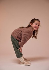 Suéter de punto rosa, pantalones bordados verdes y zapatillas beige con cintas. Niña posa de manera juguetona, mostrando elementos de un atuendo cómodo y casual.