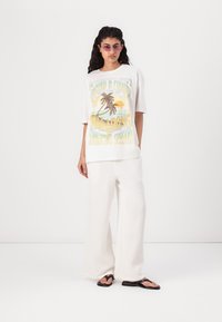 T-shirt grafico bianco con stampa da spiaggia, vestibilità rilassata; abbinato a pantaloni larghi bianchi in tessuto strutturato e ciabattine nere. Indossati occhiali da sole.
