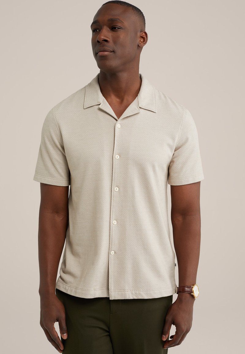 Camisa de manga corta color beige claro con un patrón texturizado, cuello abierto y cinco botones, combinada con pantalones verde oscuro y un reloj marrón.