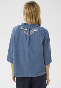 Pikkade blondide juustega naine, kes kannab sinist pluusi, millel on beežid lillekirjad selja ülaosas ja 3/4 pikkuses varrukad.