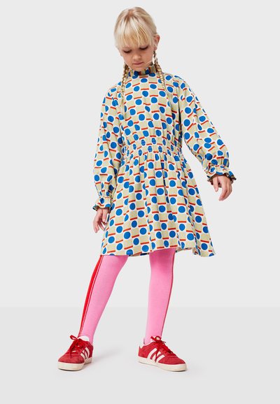 Abito arricciato con stampa colorata di cerchi blu, rossi e gialli; maniche lunghe con polsini a volant; abbinato a leggings rosa e scarpe rosse.