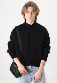 Cardigan preto de malha com decote em V, frente com botões e punhos canelados, combinado com jeans azul claro e uma bolsa cruzada preta.