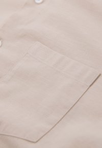 Poche de chemise beige avec coutures visibles sur un tissu léger, partie avant d'une chemise boutonnée avec des boutons blancs.