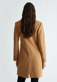 Cappotto in misto lana color cammello con una silhouette strutturata, maniche lunghe e polsini doppi, caratterizzato da una cucitura centrale e una fessura sul retro.