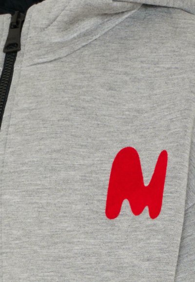 Szara rozpinana bluza z miękkiego materiału, na której z przodu po lewej stronie znajduje się duże czerwone abstrakcyjne logo. Teksturowana powierzchnia i luźny krój.