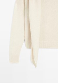 Manica di un maglione lavorato a maglia color crema con polsino a coste e un lungo nastro attaccato che pende su uno sfondo bianco.
