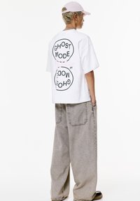 T-shirt bianco oversize con grafica testuale circolare nera "GHOST MODE" sul retro. Pantaloni larghi grigi, in materiale leggero, design semplice e senza motivi distintivi.