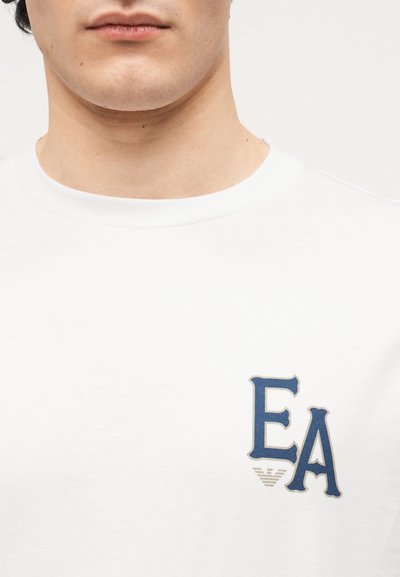 Κοντινό πλάνο ενός ατόμου που φοράει λευκό μπλουζάκι με στρογγυλή λαιμόκοψη, με μπλε γράμματα "EA" και ένα μικρό γκρι έμβλημα ακριβώς από κάτω στην αριστερή πλευρά του στήθους.