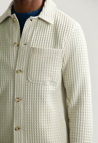 Giacca color crema, con una texture a trama quadrata, dotata di taschino sul petto, chiusura con bottoni e colletto, indossata sopra un maglione blu.