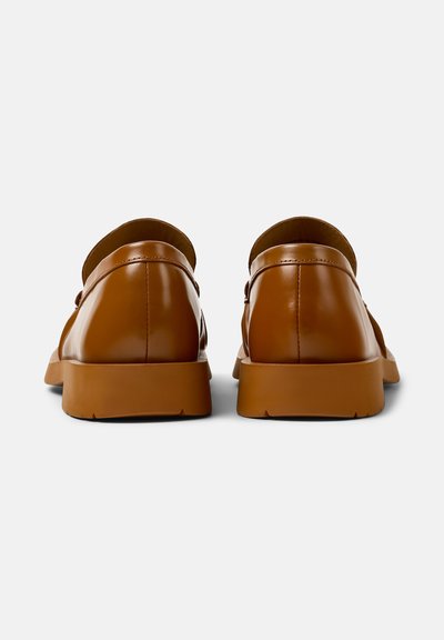 Un par de mocasines de cuero marrón pulido con suelas de goma gruesas vistos desde la parte trasera sobre un fondo blanco.