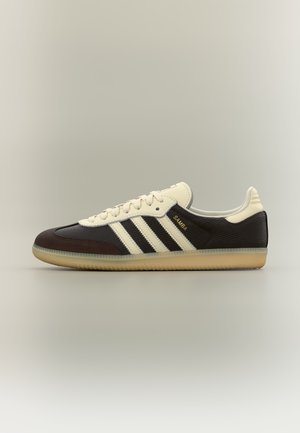 Must ja kreemjas Adidas Samba tennisking koos kummist tallaga, kreemjate paeltega ja kolme kreemja triibuga küljel, nähtav külgvaates neutraalsel taustal.