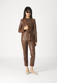 Barna kígyóbőr mintás öltöny, amely dupla soros zakót és testre szabott nadrágot tartalmaz, bézs lapos cipővel és fekete napszemüveggel párosítva.