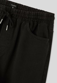 Czarne dresowe spodnie z elastycznym pasem, sznurkiem do regulacji i boczną kieszenią. Materiał posiada gładkie, teksturowane wykończenie. Widoczna metka marki.