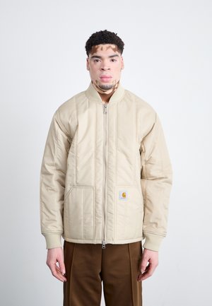 Veste matelassée beige avec fermeture éclair, poignets côtelés et poches latérales. Présente une petite étiquette logo sur la poche gauche. Texture lisse.