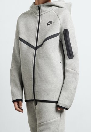 Osoba ubrana w jasnoszarą bluzę Nike na suwak z czarnym zamkiem i kieszenią na rękawie, w komplecie z dopasowanymi spodniami.