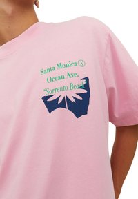 Rožinė medvilninė marškinė su mėlyna grafika, vaizduojančia lapą ir tekstu. Tekstas skelbia: "Santa Monica Ocean Ave. 'Sorrento Beach.'"