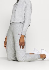 Szara przykrótka bluza z kapturem, zestawiona z dopasowanymi szarymi dresami. Dresy mają boczne kieszenie i subtelny detal z logo. Białe sneakersy dopełniają wygląd.